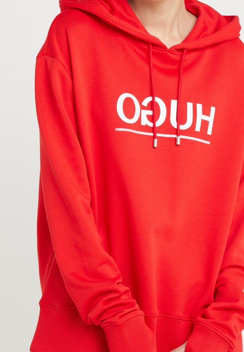 Personne portant un sweat à capuche rouge avec du texte blanc inversé "HUGO" et des cordons, montrant la partie supérieure du torse et les bras croisés.