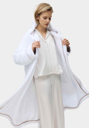 Kvinde justerer en lang hvid fluffy badekåbe over creme-stribet pyjamas med knapper og matchende bukser mod en ensfarvet lys baggrund.