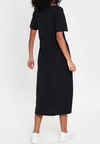 Robe noire à manches courtes de longueur maxi, confectionnée dans un tissu doux avec une taille cintrée et un design minimaliste. Portée avec des baskets blanches.