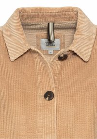 Veste en velours côtelé beige avec un col pointu, deux boutons et une boucle de suspension rayée. Texture visible et motif en côtes. Taille 36.