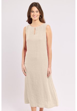Femme souriante aux longs cheveux bruns portant une robe midi beige sans manches avec une petite ouverture à l'avant et des boucles d'oreilles dorées, debout sur fond blanc.
