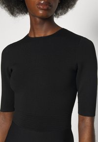 Femme à la peau foncée portant une robe noire ajustée avec des manches aux coudes et une ceinture côtelée, sur un fond blanc uni.