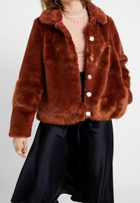 Femme portant une veste en fausse fourrure couleur rouille sur un haut rose côtelé et une jupe en satin noire, accessoirisée de bijoux et de bagues dorés.