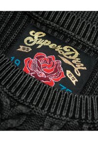 Etichetta di un maglione nero a maglia con "SuperDry" in scrittura dorata, un design di rosa rossa e accenti blu con una texture di tessuto.