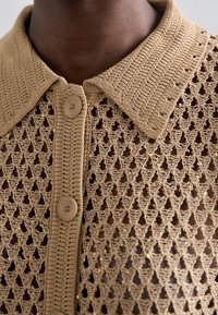 Hæklestrikket skjorte i beige farve med krave og knaplukning. Stoffet har et indviklet diamantmønster.