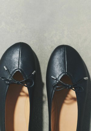Par sort sorte ballerinaer med små bundne sløjfer og runde tæer på en grå overflade.