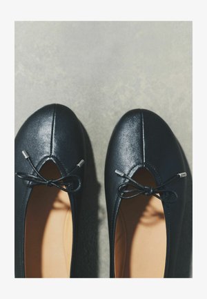 Pari mustia balettitossuja, joissa on pienet solmitut rusettinauhakuviot ja pyöreät kärjet harmaalla pinnalla.