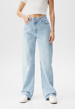Person iført lyseblå brede jeans, hvid cropped top og hvide sneakers, stående med den ene hånd i lommen på hvid baggrund.