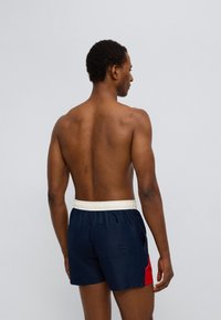 Ανδρικό μαγιό σορτς σε ναυτικό μπλε, με κόκκινο διακοσμητικό λεπτό έμφαση στο πλάι, ελαστική λευκή μέση και μία πίσω τσέπη. Ομαλή υφή υφάσματος.