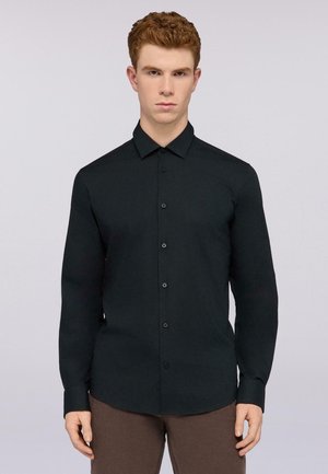 Camisa oscura de botones con mangas largas, fabricada en tejido suave. Presenta un cuello puntiagudo, seis botones frontales y un logo sutil en el pecho.