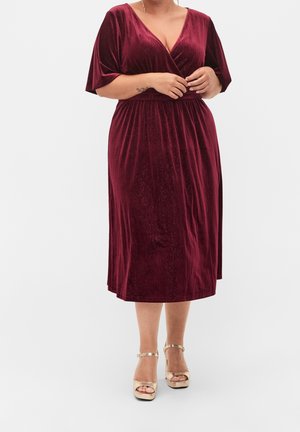 Rondingen vrouw die een knielange glinsterende bordeauxrode fluwelen jurk draagt met ellebooglange mouwen en gouden sandalen met hoge hakken.