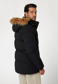 Juodas pūkinis švarkas su nuimamu dirbtiniu kailiniu gaubtu. Sukurtas su elegantišku dizainu, siuvinėtu paviršiumi ir saugiu užtrauktuku.