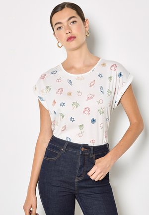 Frau trägt weißes T-Shirt mit hochgekrempelten Ärmeln, bedruckt mit bunten Motiven zum Thema Meer, und dunkelblaue Jeans mit hoher Taille, posiert mit einer Hand in der Hüfte.