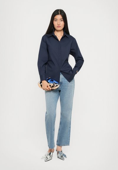 Camicia azzurra con bottoni navy a maniche lunghe, abbinata a jeans dritti azzurri chiari e mocassini argentati con dettagli a fiocco. Accessoria con una pochette blu.