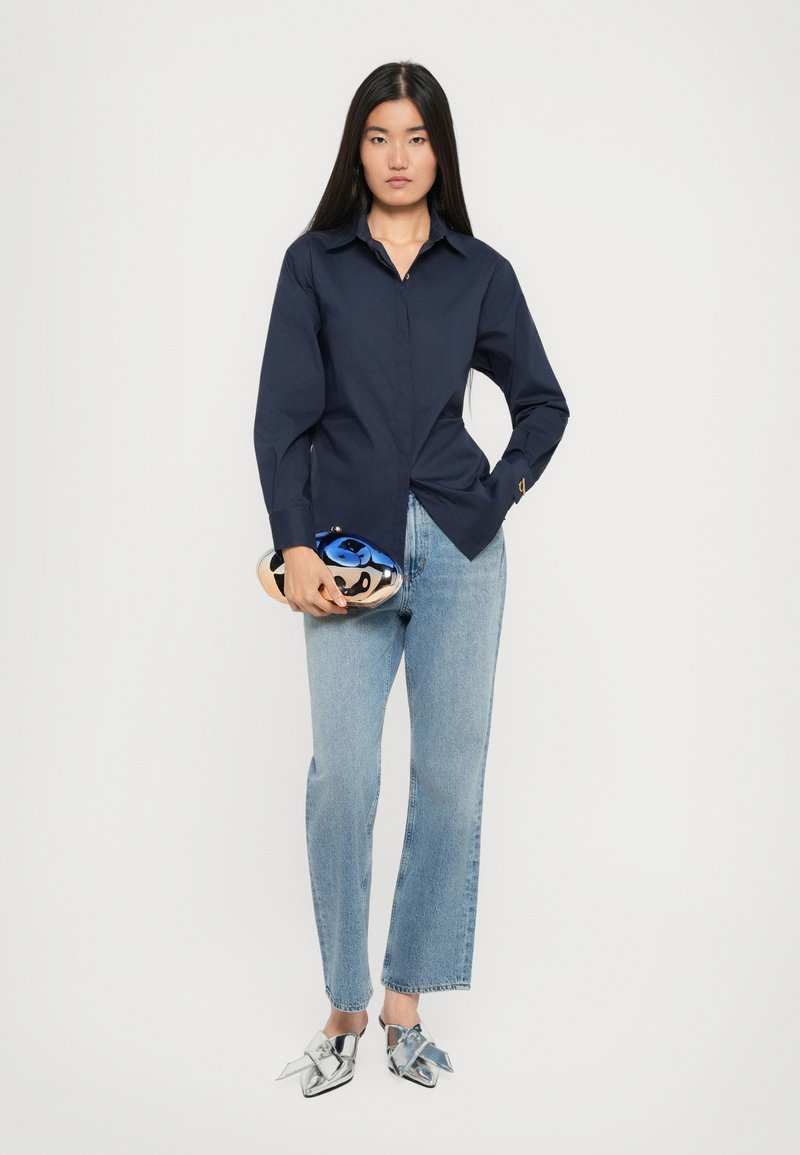 Chemise bleu marine à boutons avec de longues manches, associée à un jean droit bleu clair et des mocassins argentés avec un détail en nœud. Accessoirisée d'une pochette bleue.