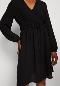 Schwarzes Kleid mit langen Ärmeln, V-Ausschnitt, gerafftem Taillenbereich und Kordelzugdetails. Hergestellt aus leichtem Stoff mit weicher Textur.