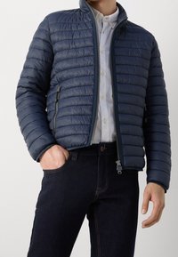 Marineblaue Steppjacke mit hohem Kragen, gestepptem Design, Frontreißverschluss und Seitentaschen, kombiniert mit dunkelblauen Jeans.