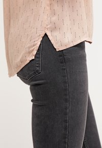 Chemise beige avec de petites lignes noires légèrement rentrées dans un jean noir, montrant la couture latérale et le détail de la poche.