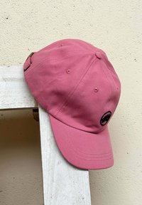 Gorra de algodón rosa con logotipo negro, diseño estructurado, visera curvada y correa ajustable con hebilla, reposando sobre una superficie de madera blanca.