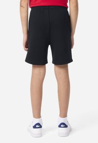 Shorts noirs avec une ceinture élastique, texture lisse, longueur au genou, associés à des baskets blanches avec un détail de logo bleu et vert.