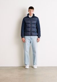 Σακάκι πούφ με ναυτικό χρώμα, οριζόντιες ραφές, κλείσιμο με φερμουάρ και κουκούλα, συνδυασμένο με ανοιχτό μπλε τζιν και λευκά αθλητικά παπούτσια. Λείος υφή.