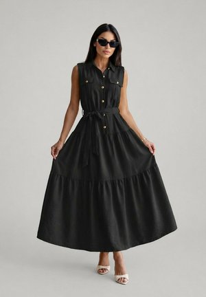 Femme portant une robe maxi noire sans manches boutonnée sur le devant avec ceinture, lunettes de soleil noires et talons ouverts blancs, debout devant un fond gris.