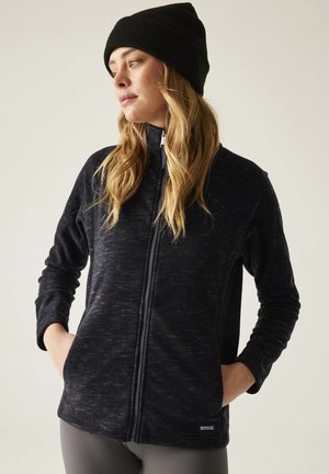 Kobieta w czarnej dzianinowej czapce i ciemnej kurtce na zamek, stojąca z rękami w kieszeniach, patrząca w lewo na jednolitym tle.