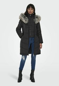 Sort puffjakke med en foret hætte, lynlåslukning og quiltet design. Bæret over blå slidte jeans og sorte hælstøvler.