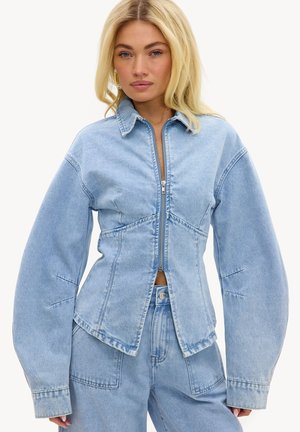 Mujer rubia con chaqueta vaquera azul claro sobredimensionada con mangas anchas y vaqueros de talle alto, de pie frente a un fondo liso.