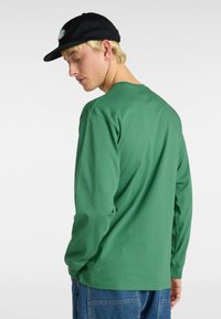 Grünes Langarmshirt aus Baumwolle mit Rundhalsausschnitt und lockerer Passform. Kombiniert mit blauen Jeanshosen und einer schwarzen Mütze.