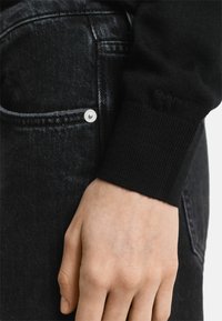 Zwarte trui met geribbelde manchetten, gecombineerd met donkere jeans die een zilveren knop en zakdetails hebben. De gladde stoftextuur is zichtbaar.