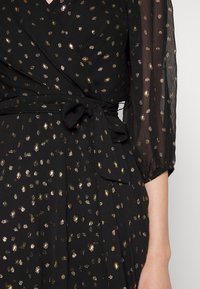 Robe noire avec un motif à pois dorés, design portefeuille et détail noué à la taille. Manches bouffantes transparentes avec poignets élastiques. Tissu doux et texturé.