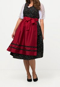 Femme portant une robe noire à petits motifs floraux, manches courtes en dentelle blanche, avec un tablier rouge noué sur la jupe, accompagnée de talons noirs.