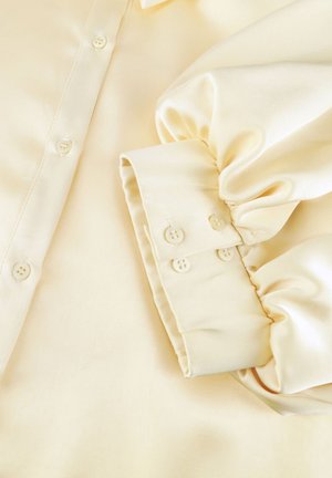 Crèmekleurige satijnen blouse met lange pofmouwen, knoopmanchetten en een knoopsluiting aan de voorkant.