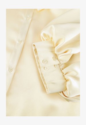 Crèmekleurige satijnen blouse met lange pofmouwen, knoopmanchetten en een knoopsluiting aan de voorkant.