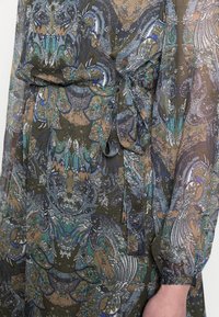 Průhledné šaty s vzorem v zemitých tónech a vířivými květinovými motivy. Obsahují pásek v pase a dlouhé rukávy se staženými manžetami.