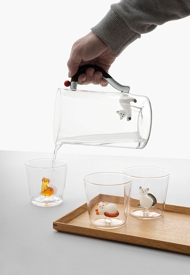 Przezroczysta szklana dzbanek z czarnym uchwytem, nalewający wodę do trzech przezroczystych kubków z motywami zwierzęcymi. Pod kubkami znajduje się drewniana taca.
