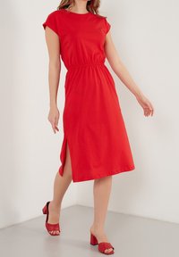 Rotes, kurzärmliges Jerseykleid mit elastischem Bund und Seitenschlitz. Kombiniert mit passenden roten Absatzsandalen auf einem hellen Hintergrund.