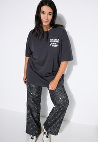Czarnoszary, oversize t-shirt z białym nadrukiem hasła, w zestawie z czarnymi błyszczącymi spodniami typu dzwony ozdobionymi srebrnymi akcentami oraz białymi sneakermi.