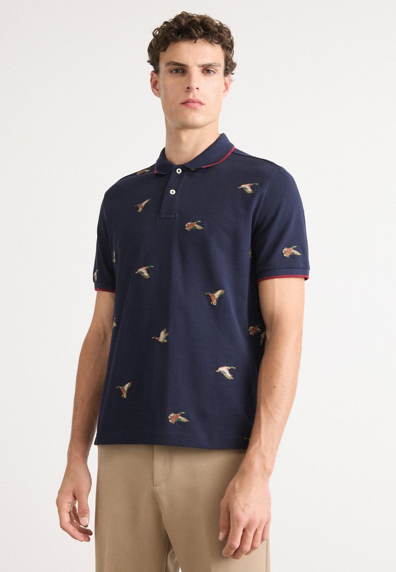 Marineblå polo shirt laget av bomull, med broderte andemotiver, en rød krage og korte ermer. Glatt tekstur, knappestolpe.