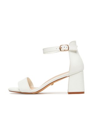 Bílá sandála s otevřenou špičkou, širokým podpatkem a nastavitelným páskem na kotníku se zlatou sponou, zobrazena z bočního profilu na bílém pozadí.