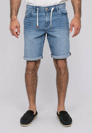 Homme portant un short en jean bleu clair retroussé avec un cordon de serrage blanc et des chaussures noires sans lacets, debout les bras détendus le long du corps.