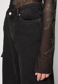 Κοντινό πλάνο μαύρων τζιν με χάλκινες ραφές και μια μαύρη διάφανη μπλούζα με μακριά μανίκια που διαθέτει σχέδια από ρουμπίνι στο μανίκι.