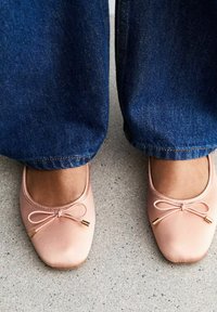 Ljust rosa balettskor med rund tå, prydda med en dekorativ rosett och guldaccenter, kombinerade med blå denimjeans. Betongytan nedan.