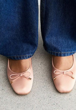 Light pink ballet flats with a rounded toe, featuring a decorative bow and gold accents, paired with blue denim jeans. Concrete surface below.