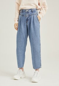 Fark yüksek bel, açık mavi denim, plise detaylı, dar paçalı ve klasik düğme kapamalı. Beyaz spor ayakkabılar ve açık pembe bir üst ile kombinlenmiş.