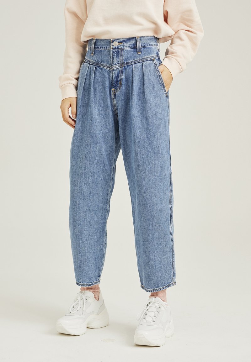 Fark yüksek bel, açık mavi denim, plise detaylı, dar paçalı ve klasik düğme kapamalı. Beyaz spor ayakkabılar ve açık pembe bir üst ile kombinlenmiş.