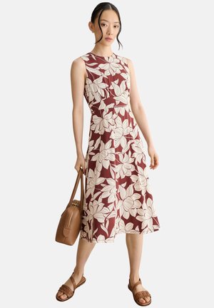Mujer con vestido sin mangas color burdeos con gran estampado floral blanco, sosteniendo un bolso marrón y calzando sandalias marrones.