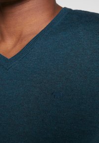 Maglione lavorato a maglia color verde petrolio scuro con scollo a V, caratterizzato da un logo ricamato in modo sottile sopra il lato sinistro del petto. Tessuto strutturato con collo a costine.