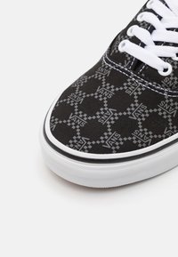 Pointe de chaussure Vans noire avec des lacets blancs et un motif à carreaux répétitif gris du logo Vans sur la tige en tissu.